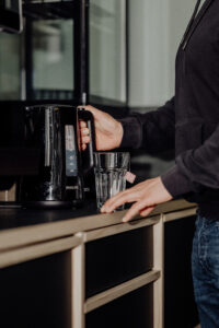 Eine männliche Hand an einem Wasserkocher. Vor dem Gerät steht ein Glas mit Teebeutel.